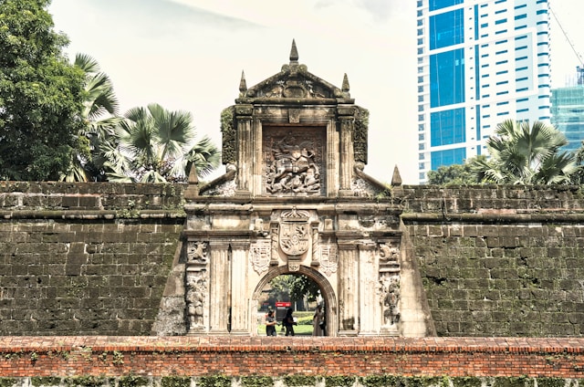 Historic stone gate of Intramuros with detailed carvings, lush palm trees in the background, and a modern skyscraper to the right, blending eras.